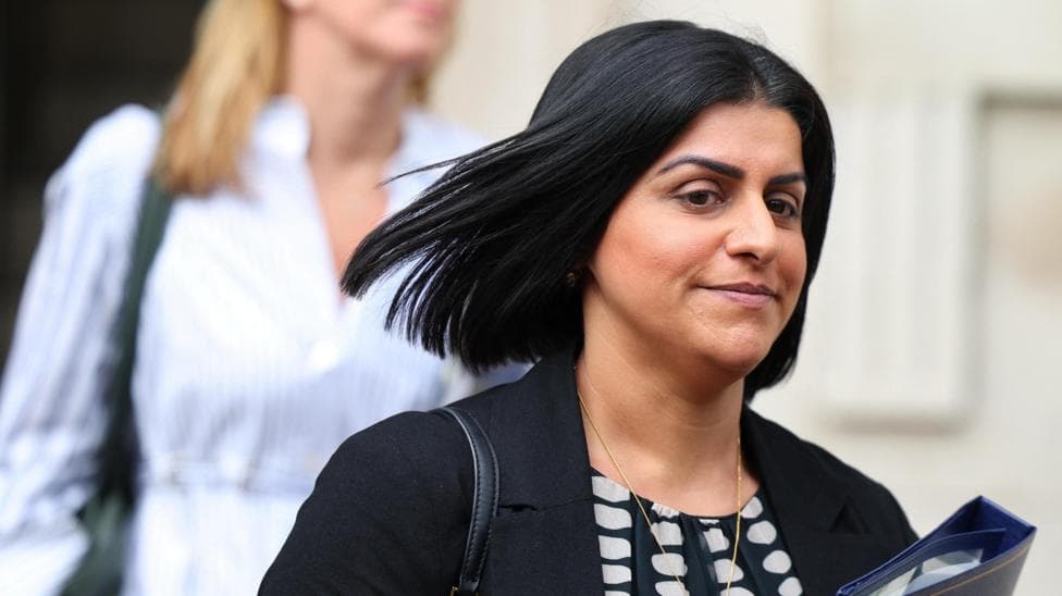 Shabana Mahmood is holding a blue folder. She is wearing a black jacket, a black and white top, and has a bag strap over her right shoulder.
