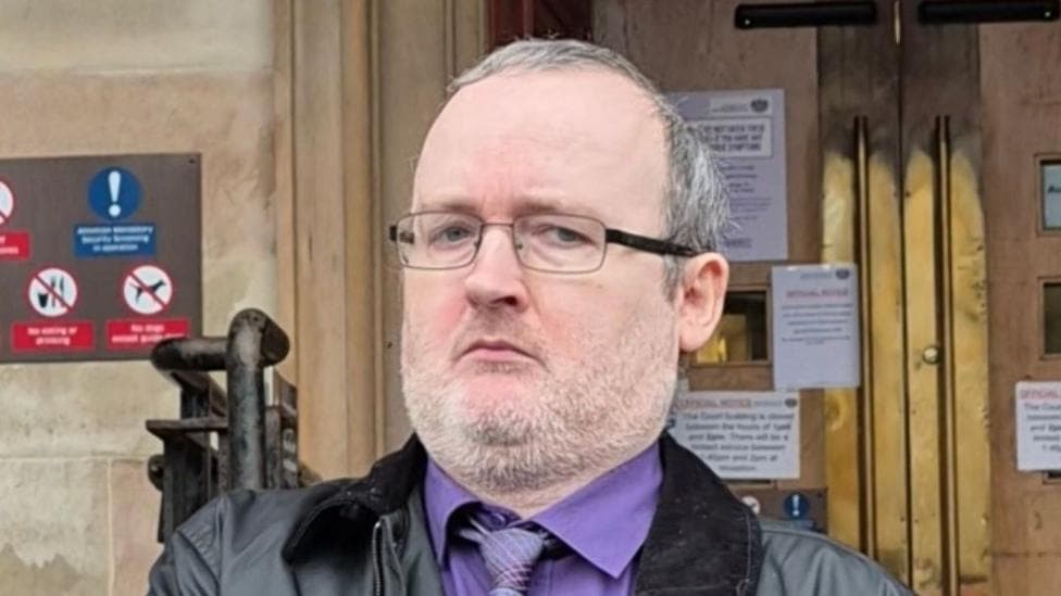 Martin Gallacher leaving court. He has glasses, short greying hair and a thin beard with white and grey hairs in it. He has a purple shirt and dark jacket on.