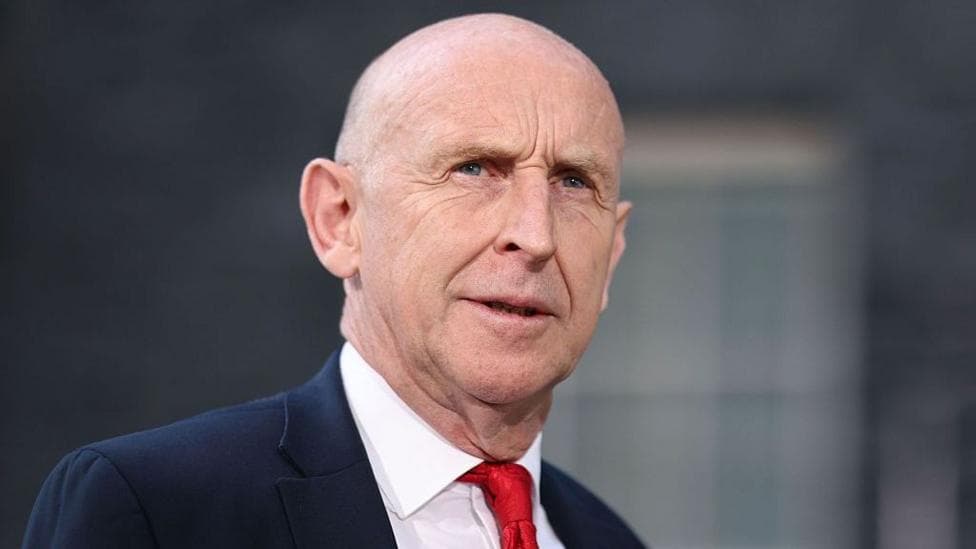 John Healey stands looking into the middle distance wearing a red tie, white shirt and navy blue suit. There is a blurred window behind him.