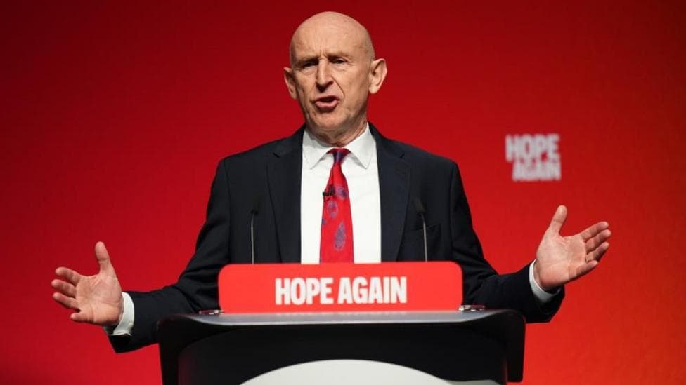 Johm Healey has his arms out in a gesture while speaking at a podium in front of a red backdrop.