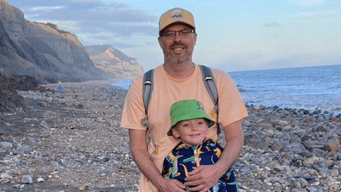 Jonathan and his young son stand together on a rocky beach, with Jonathan in a light orange t shirt with his arms around the boy in a dinosaur pattern blue wind breaker and green bucket hat. Steep cliffs stretch along the shoreline behind them, with the sea to their right.