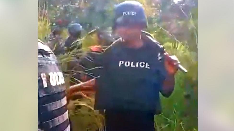 A screen grab from a video shows a Nigerian police officer wearing a navy bulletproof vest and a a helmet while walking through a forest. Several other officers can be seen in the background.