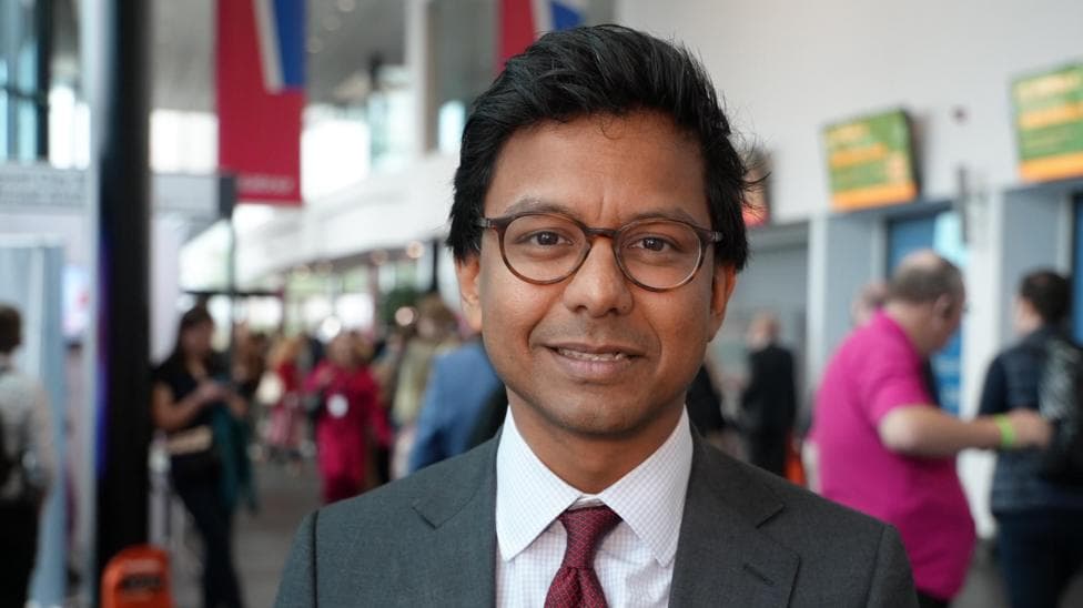 Kanishka Narayan, wearning dark rimmed glasses and a suit and tie, poses for the camera at a party conference