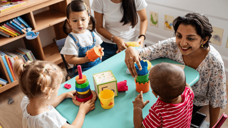 A group of very young children around a table, with a teacher. They have an assortment of toys. A bookshelf can be seen in the background.