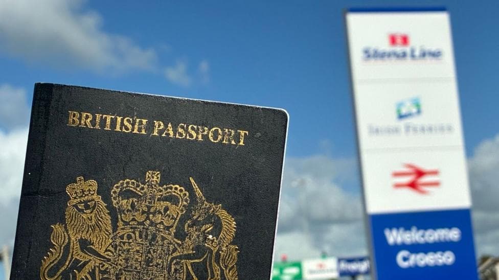 A dark blue British passport in the foreground with a sign welcoming people to Holyhead port, slightly out of focus in the background