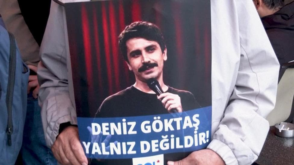 A poster of Deniz Göktaş, who has dark short hair and a moustache, holding a microphone with a red curtain behind him. The words "Deniz Göktaş is not alone!" are written on the poster.