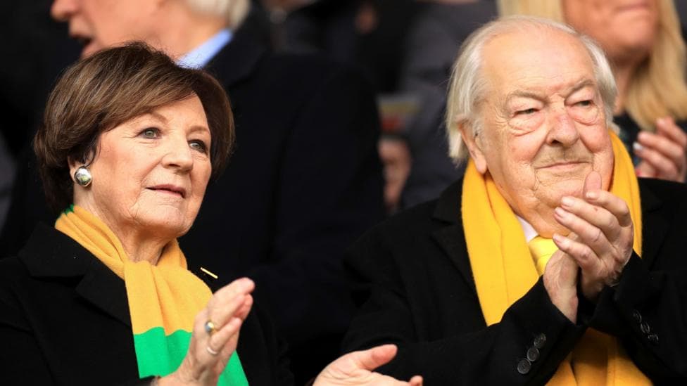 Delia Smith and Michael Wynn Jones applauding in the stands at a Norwich City football match. They are both wearing black outfits with green and yellow scarves around their necks.
