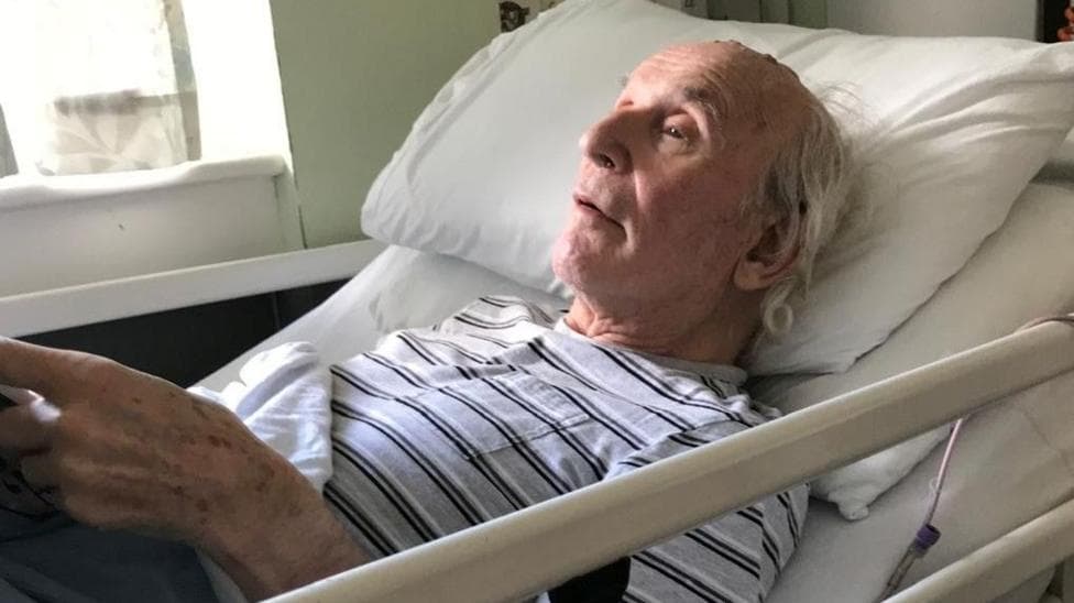 John Russell is an older man. he is wearing a black and white stripy T'shirt and lying back in a hospital bed.