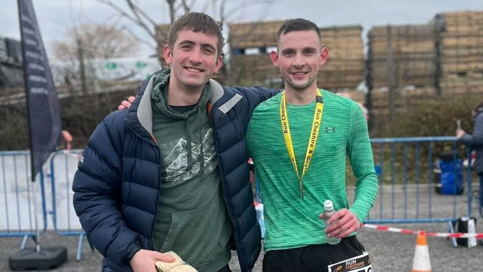 The two men have their arms around each other. The one on the left has brown hair and is wearing a green hoody and puffer jacket. The man on the right is wearing a bright green sports top and has a medal around his neck and he is holding a bottle of water. There are barriers in the background.