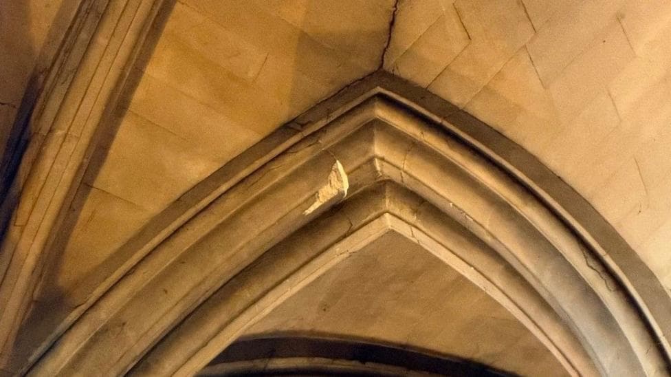 The photo shows a close-up of the high ceiling in the south nave of Bristol Cathedral. An intricately carved stone arch can be seen, with a chunk of stone missing. The damaged area looks paler than the rest of the arch. There is also a crack in the ceiling above the arch itself.
