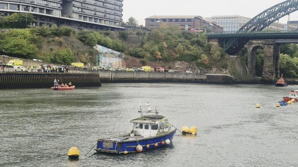 Ambulances, police cars and a fire engine line the river bank in Sunderland by an orange RNLI rescue boat. Two small fishing boats are also in the River Wear. The image is taken from the opposite bank.