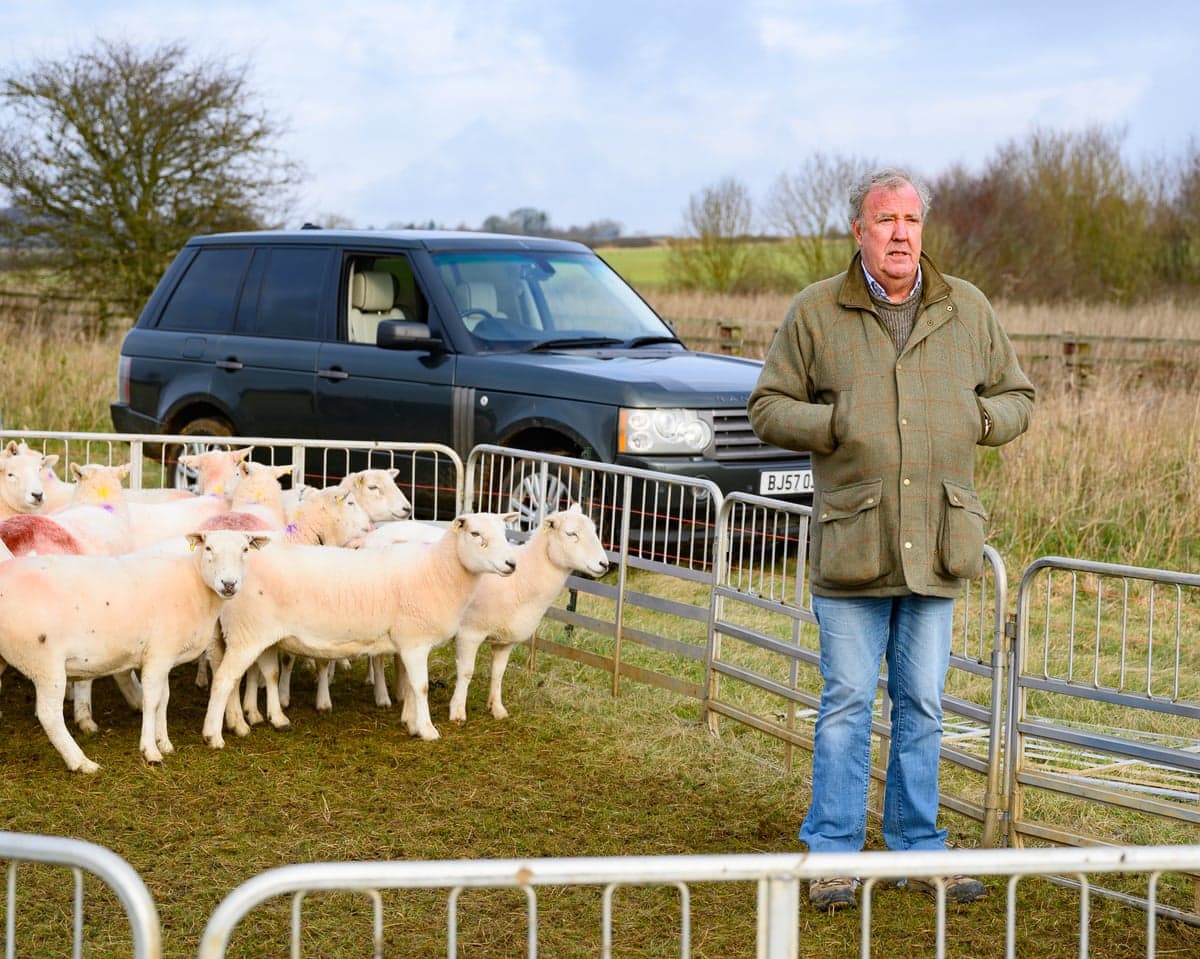 Jeremy Clarkson stands near a fence, with sheep and a black car behind him