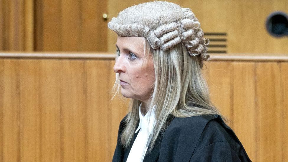 A woman with blonde hair, wearing black robes and a traditional court wig, in profile