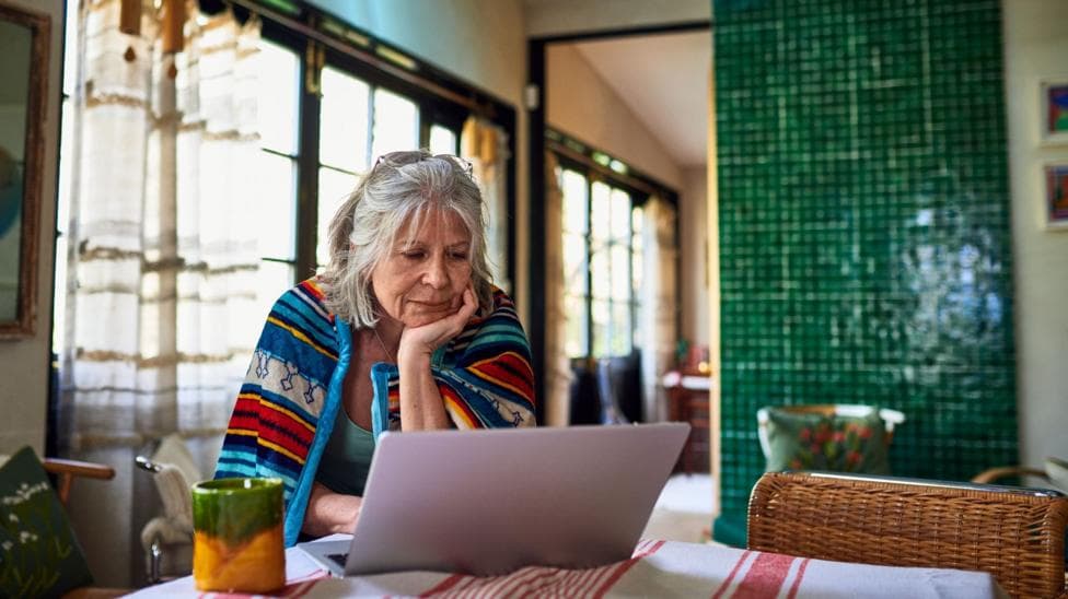 A pensioner looks at her finances on her laptop at her dining room table. She is wearing a colourful throwover.