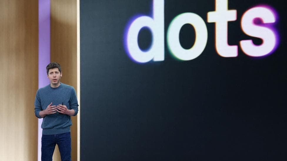OpenAI CEO Sam Altman standing on stage at his company's annual developer conference in front of a screen with "dots" displayed behind it.