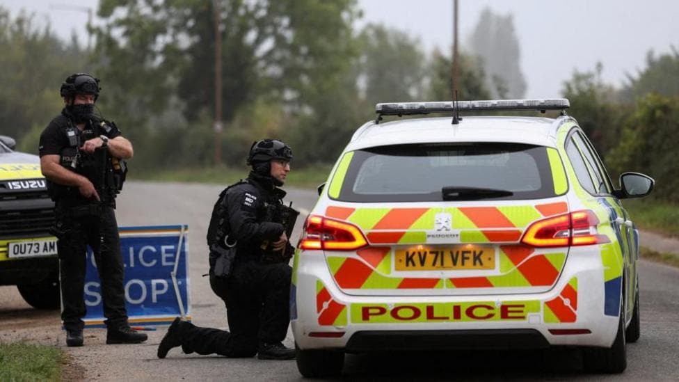 An armed officer stands in uniform as another one crouches down with a gun in his hand pointed down and he talks through the window of a police car which has its back with a sign 'police' visible