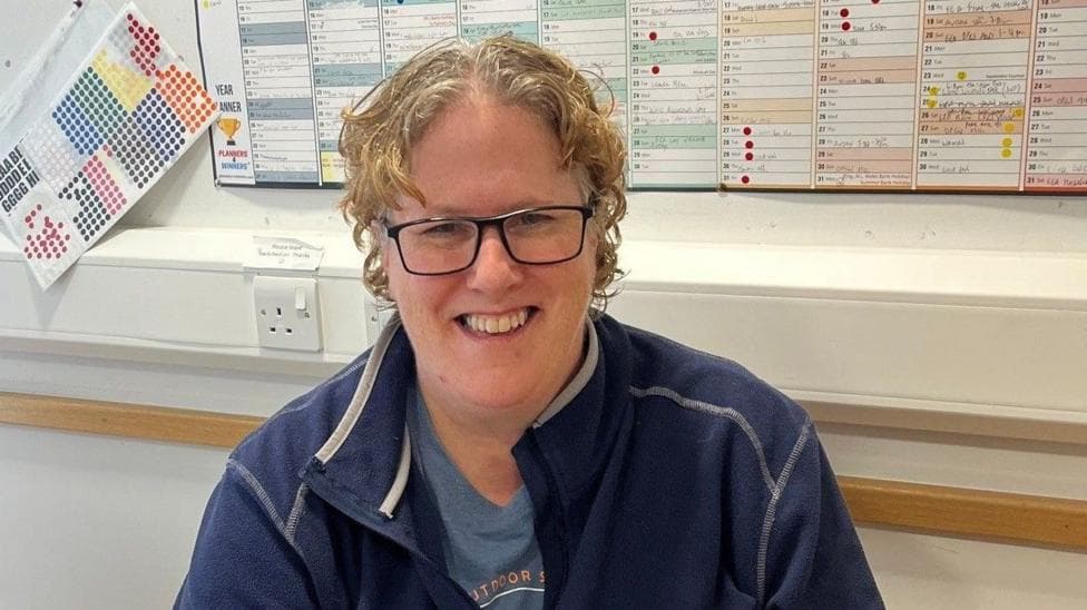 Trudi, a woman with curly blonde hair and glasses, sits at a desk in a blue zip-top with a blue T-shirt underneath. There is a wall planner behind her and plug points.