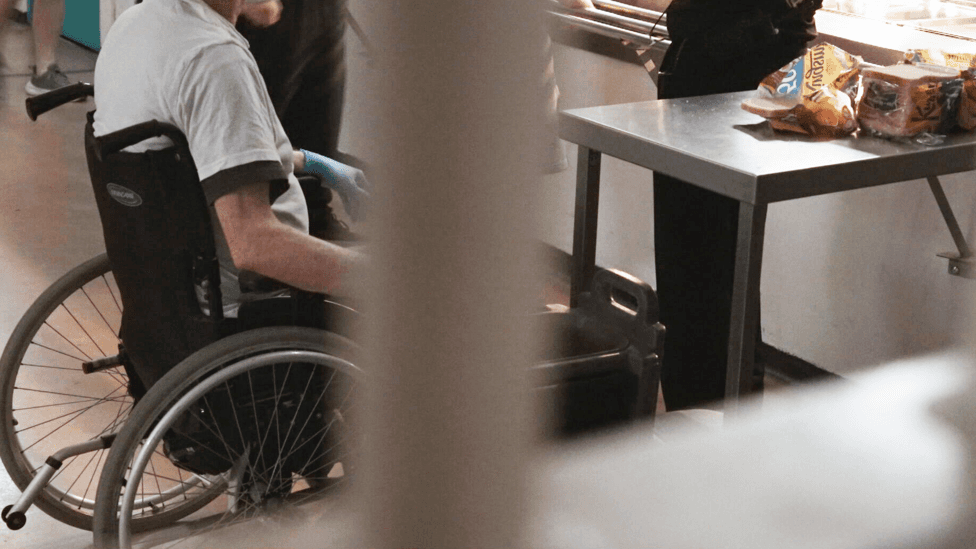 Inmate in a wheelchair. You cannot see the man's head. He is in a room behind bars where food is being served