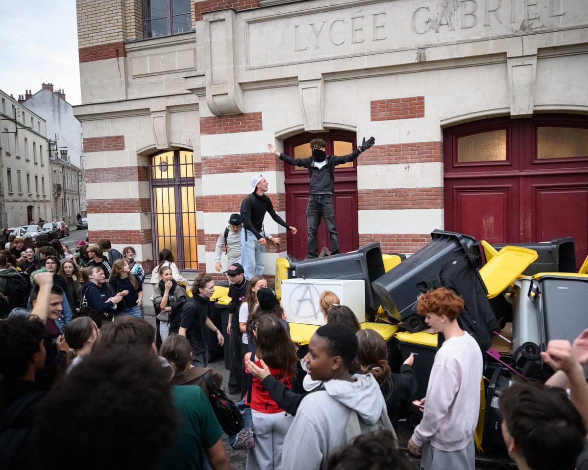 A masked protester stands on piled bins outside a secondary school during a student blocking protest