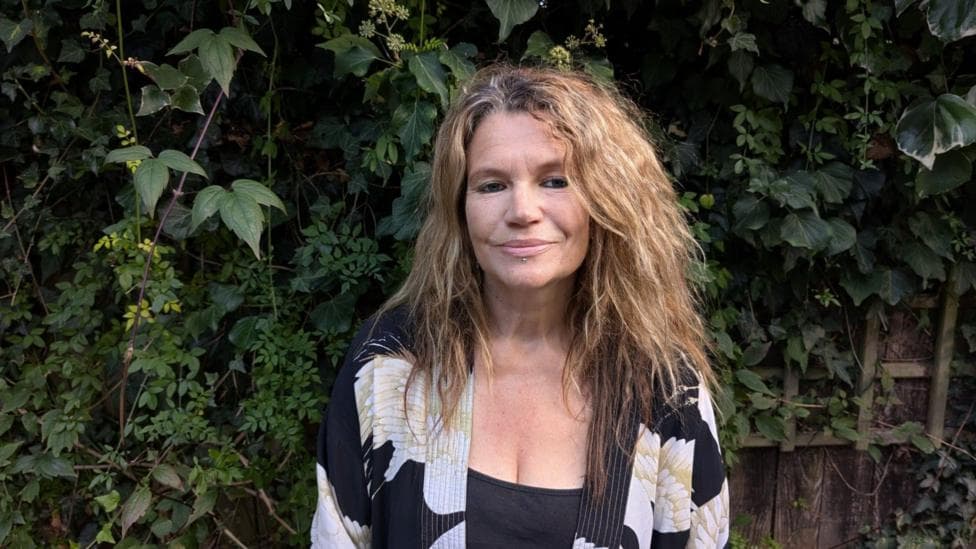 Helen with shoulder length wavy brown hair wearing a black vest and a black white and gold kimono style top standing in a garden with fence and foliage behind her