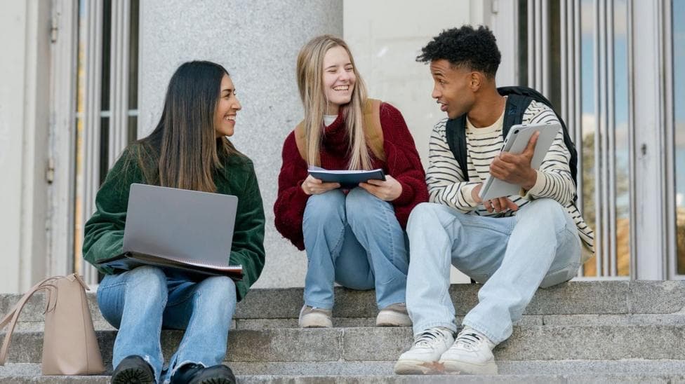 Three university students sitting on concrete stairs, talking and smiling while looking at a digital tablet and laptop. There are two women to the left both looking at a male student to the right.