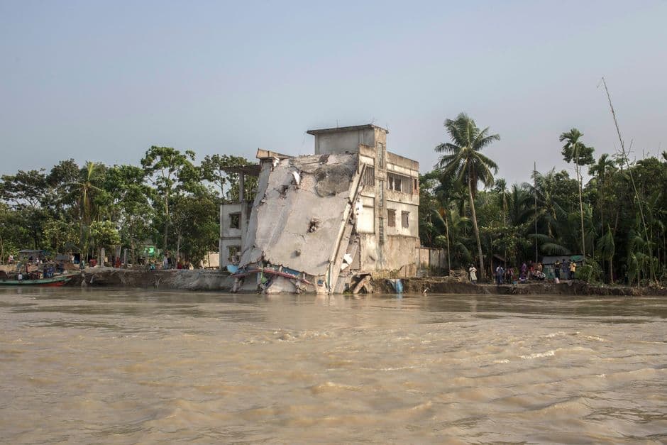 A damaged building collapsing by a tropical riverbank with palm trees and vegetation.