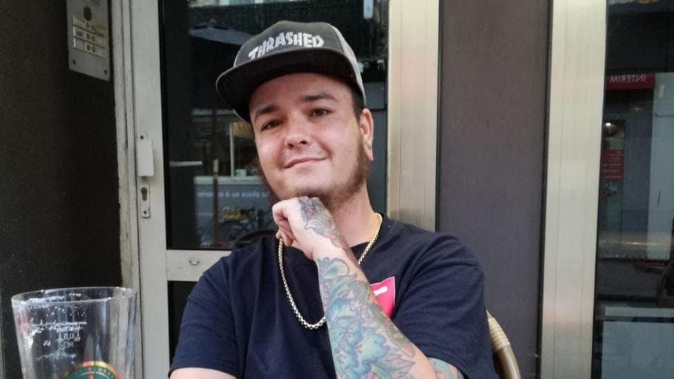 Hobie Harrison looks at the camera and smiles. He is sitting at a table with what's left of a beer. He looks happy and has a beard. He has a thin gold chain around his neck and a black T-shirt. He has a baseball cap on.