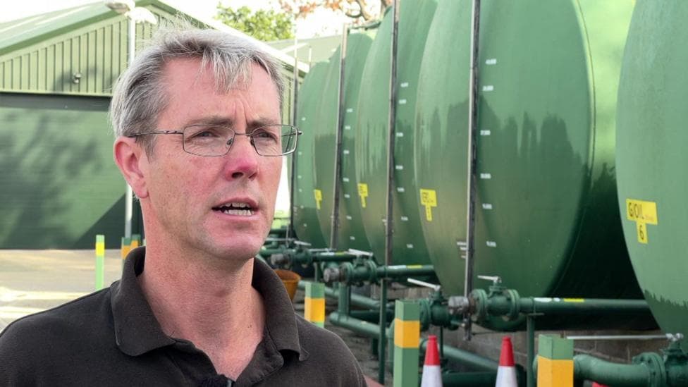 Dave Marsh stands next to a row of large, green oil tanks as he gives a TV interview. He has a company Polo shirt, grey hair and glasses.