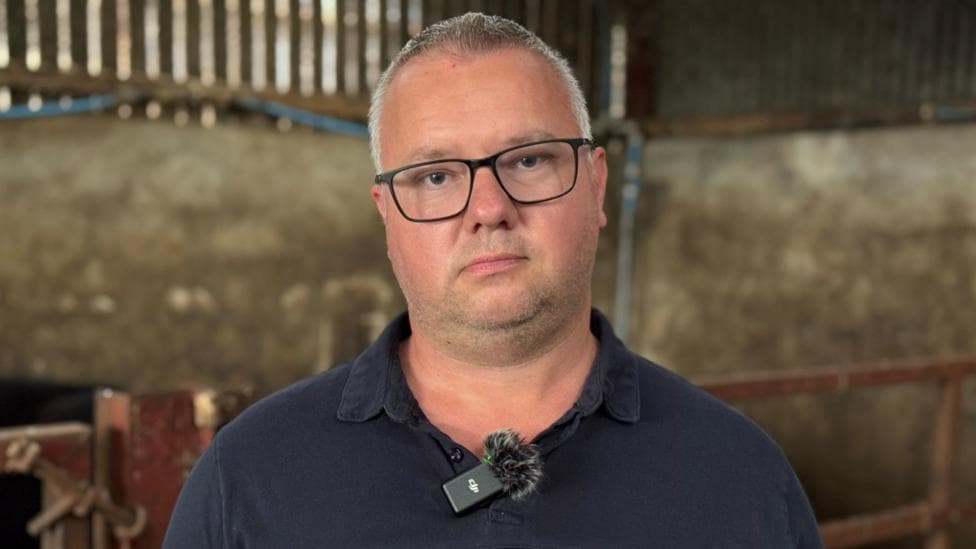 A man with short white hair looks into the camera. He has dark stubble and is wearing square glasses. He is wearing a navy polo shirt and has a microphone attached to it. He is standing in a barn.
