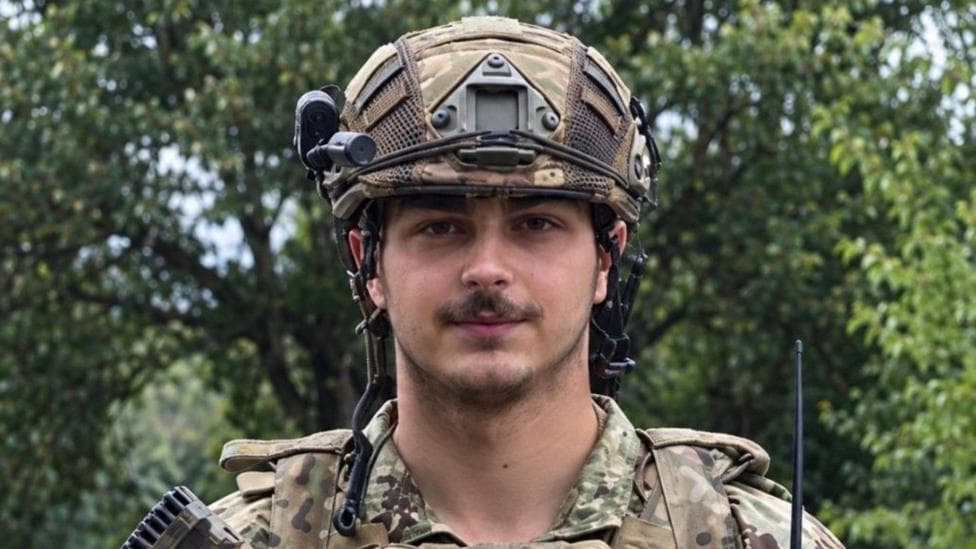 A young soldier with a moustache in a military helmet and army uniform with trees behind him