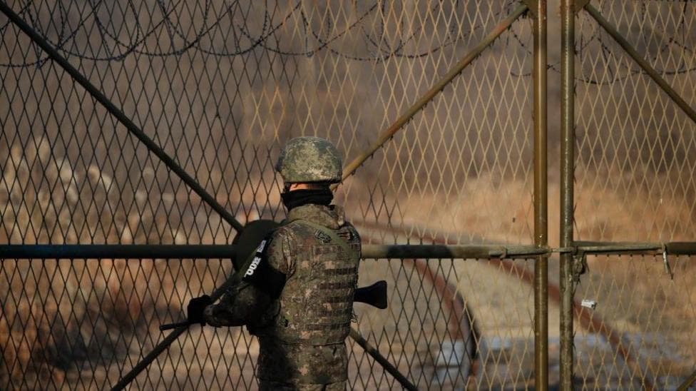 A soldier in camouflage uniform and helmet stands guard in front of a tall metal fence topped with barbed wire. Beyond the fence, a narrow railway track and barren winter landscape stretch into the distance.