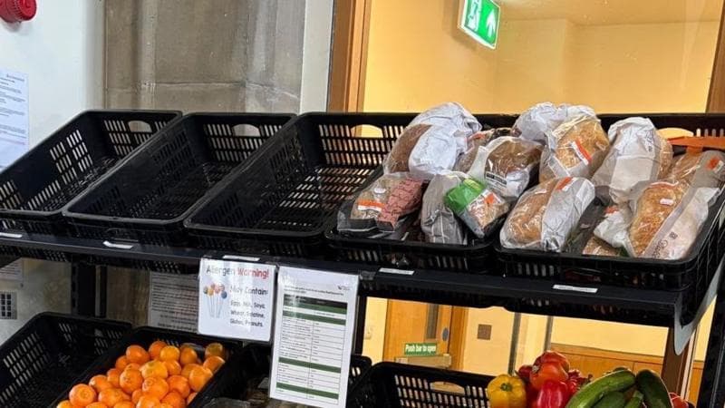 Shelving in a community food pantry displays packaged loaves of bread on the top shelf, with empty black baskets beside them. Below, signs providing allergy and food information hang above trays of fruit and vegetables, including oranges, peppers, and courgettes. The display is situated indoors beside a glass wall and a lit emergency exit sign.