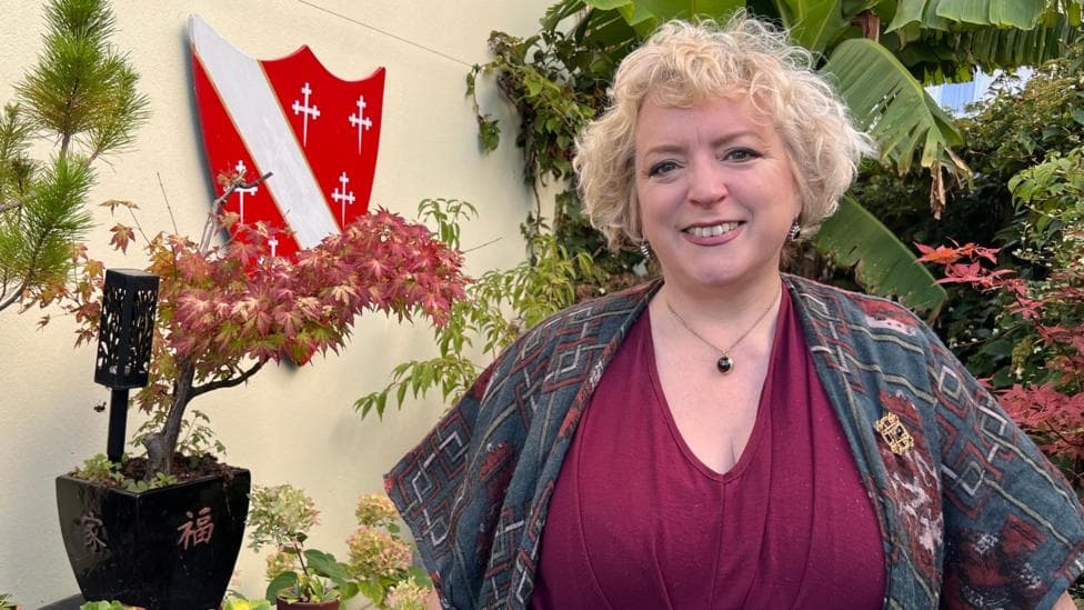 Charlotte Howard in a busy garden smiling at the camera, she has short blond hair and wears a maroon V neck top and blue and red shawl.