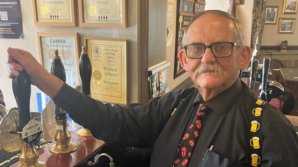 Glyn Jones, a man standing behind the bar in The Prince Albert pub in Ely, Cambridgeshire. Glyn is ready to pull a pint and is looking directly at the camera. He is wearing a black shirt with a novelty tie which has mouths on and a pair of braces which has pints on it.