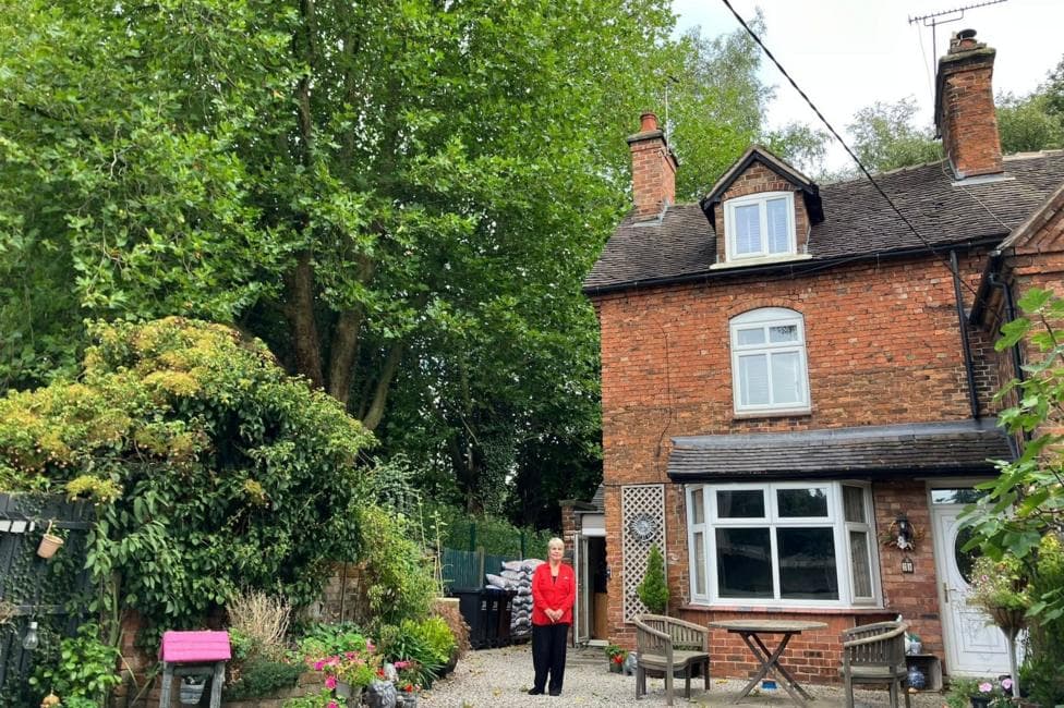 Maureen Forsyth has blonde hair and is standing in her garden. She is wearing a red top and black trousers and standing with her house behind her, which has a large tall tree very close to her house and branches overhanging her garden. Some are less than a metre away from the chimney