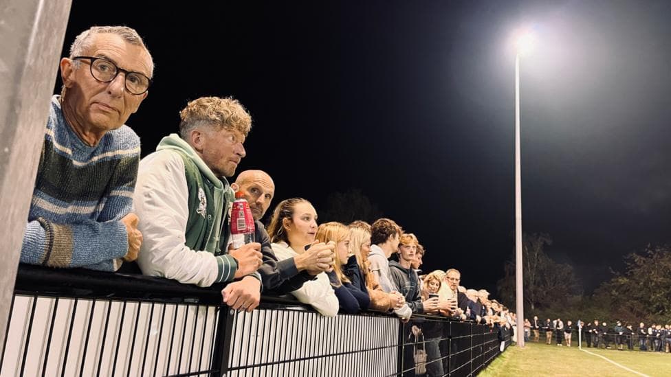 Fans stand behind a fence looking out at the pitch where the match is being played