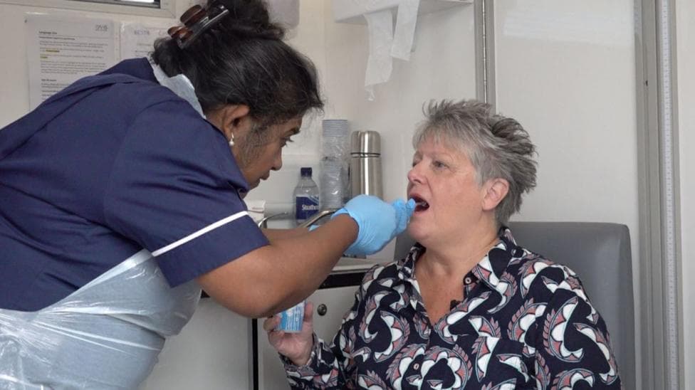 A woman sits in a chair with her mouth open, while a nurse in scrubs and protective clothing including gloves and an apron inserts a pill into her mouth.