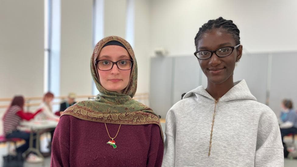Two teenage girls look directly at the camera. One is wearing a grey hoodie and the other is wearing purple jumper and a headscarf.
