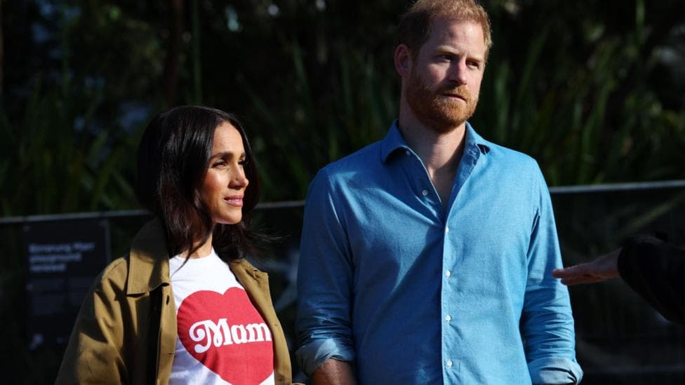Harry and Meghan seen walking together in Sydney harbour in April. He is wearing a blue shirt and a cap, she is wearing a white tshirt emblazoned with a love heart and a tan coloured jacket.