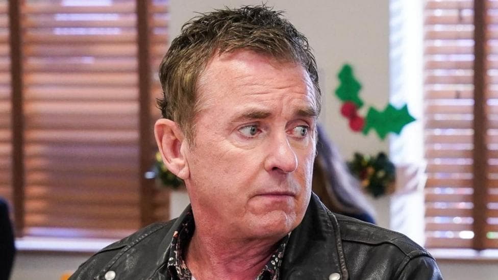 Actor Shane Richie as Alfie Moon in EastEnders. A head and shoulders picture of him sitting in a room looking to his left. He is wearing a leather jacket with a floral shirt underneath.