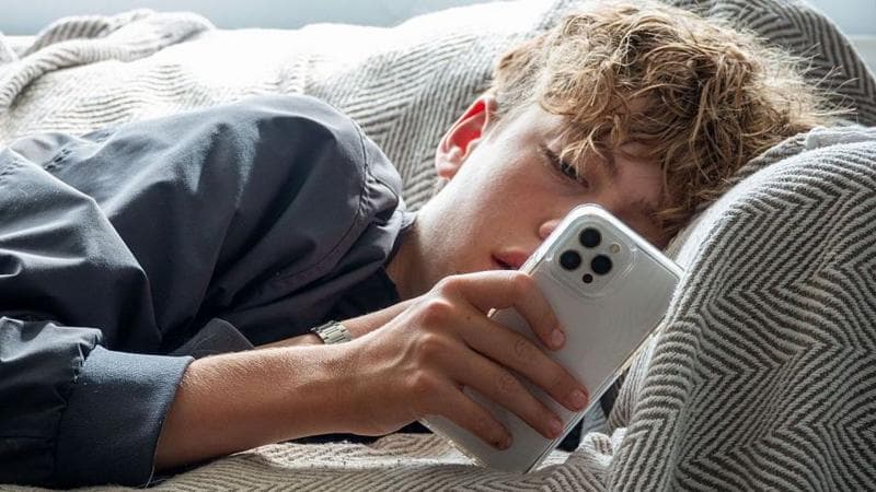 Young person with curly brown hair lying on a sofa, looking at a white phone
