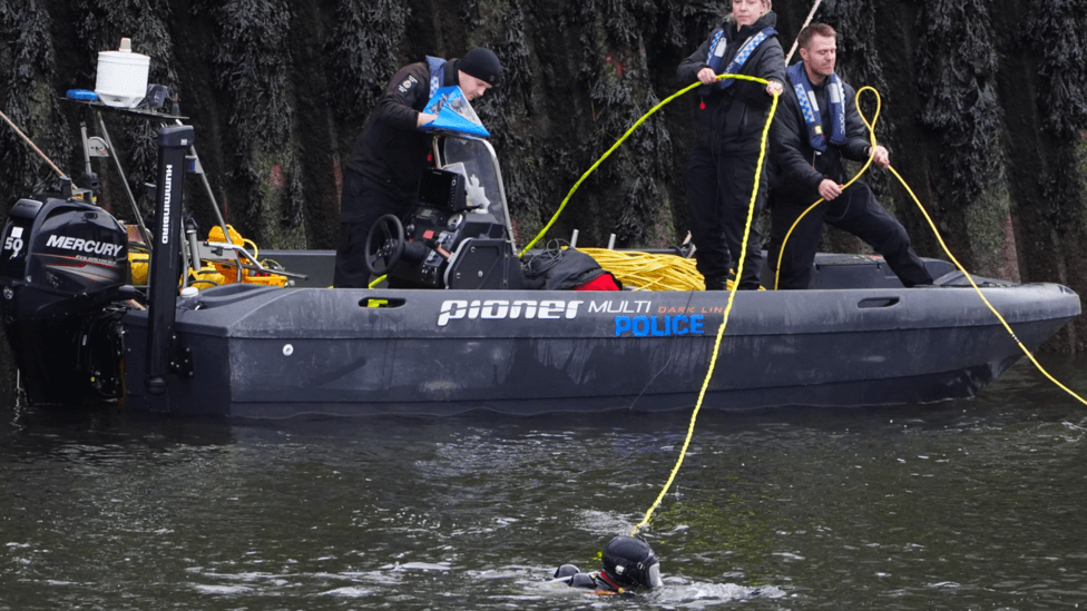 Police officers and divers - wearing black uniform - stand in a rubber dinghy RIB over the River Wear. There is a diver in the water who has been linked to the boat via a bright yellow safety line. They are searching that.