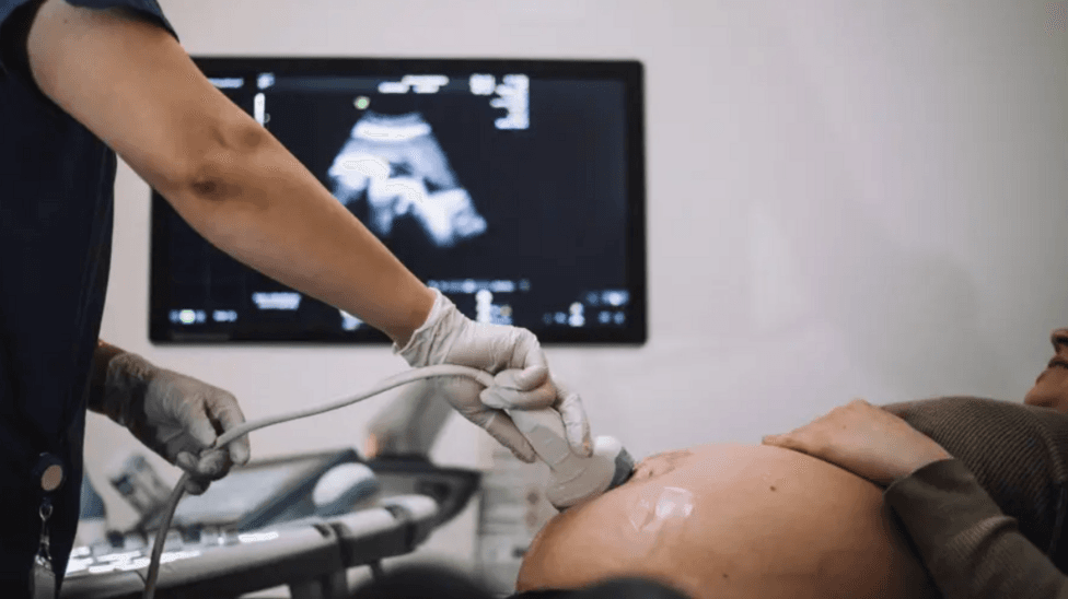 A woman is undergoing a pregnancy scan in hospital - her pregnant stomach is visible, and the arm of the medic, with a screen behind them
