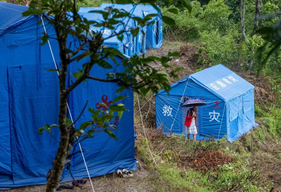 Blue disaster-relief tents on a muddy, forested hillside. A person holding an umbrella stands outside one of the tents. White Chinese characters and a relief-aid logo are visible on the bright blue tent walls.