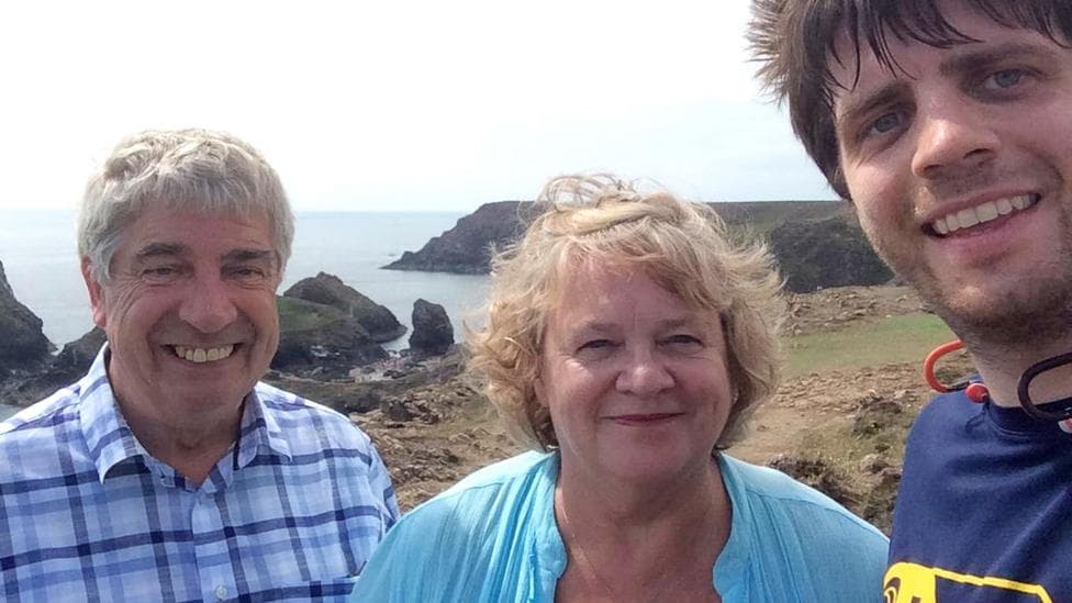 An older woman with light blonde hair and a bright blue t-shirt smiles at the camera and poses next to a younger man, also smiling, who seems to be taking the photo as a selfie. Next to the woman on the left is a man grinning and showing his teeth, wearing a blue chequered shirt. They are standing on a clifftop walk with coastline in the background.