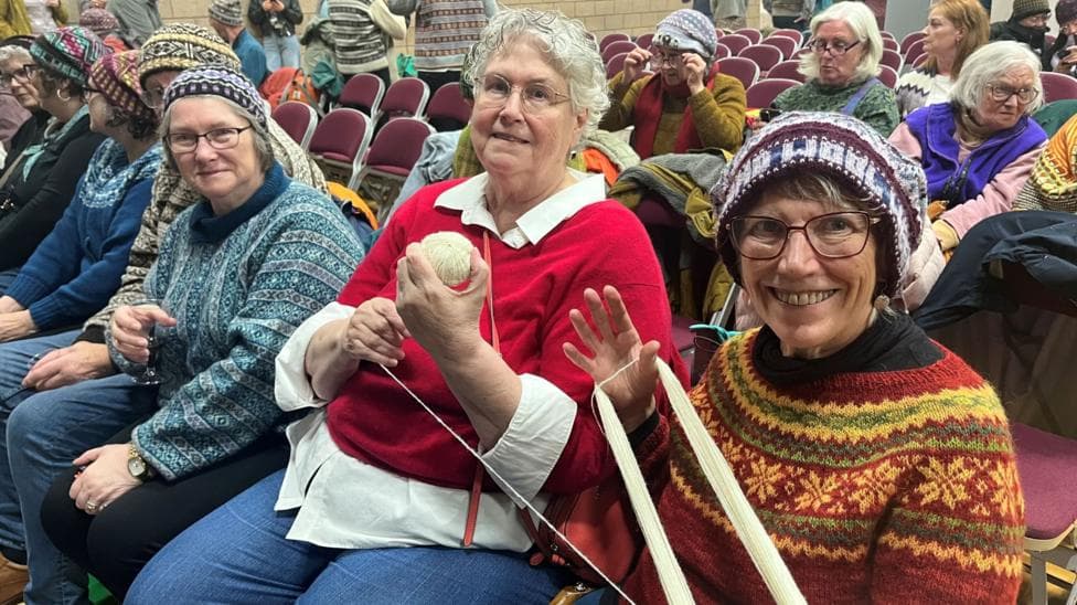 A group of female knitters are sitting in chairs in a large hall. Some of them are wearing woollen hats, while the two women nearest the camera are working with a ball of wool.