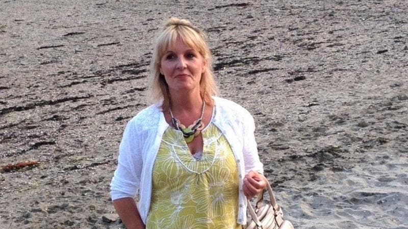 A woman with blonde hair standing on a beach wearing a yellow dress and a white cardigan