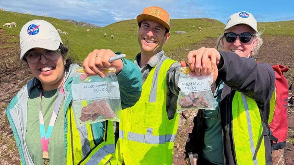 The three scientists are standing in a flat landscape of rocks and grass. They are wearing caps and smiling as they hold up sample bags containing rock samples.