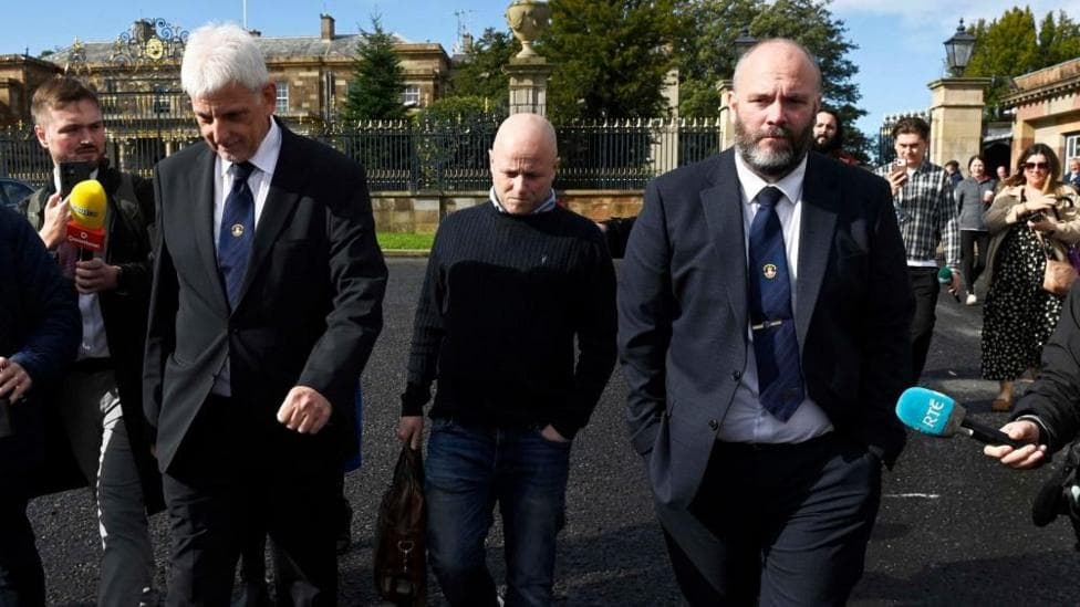 Three men walk side by side as members of the media point microphones in their direction. Two of the men are wearing dark suits, white shirts and navy ties. The other man is wearing a navy jumper and blue jeans.