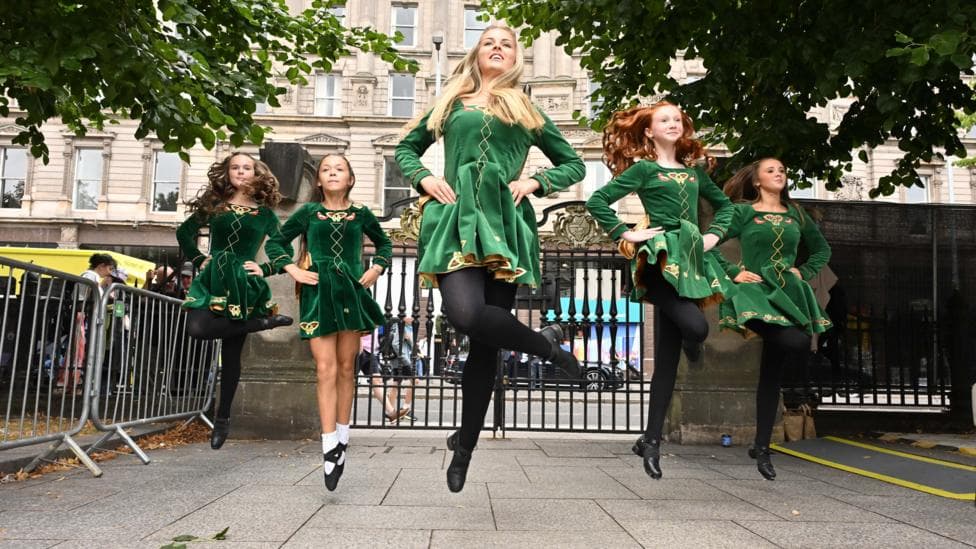 A woman and four girls are Irish dancing. The woman is in the centre with two girls on each side of her. They're wearing green dresses and black dance shoes. All are propelled off the ground.